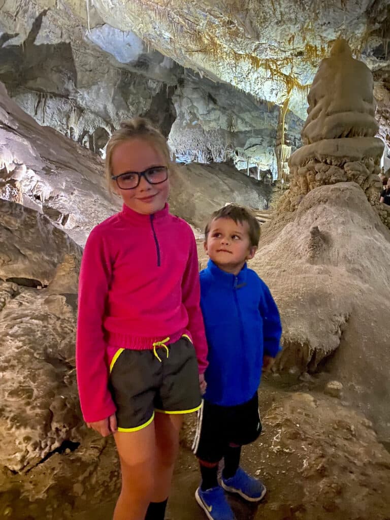Two young children, a girl in a bright pink jacket and a boy in blue, stand inside a cave with rocky walls. The scene captures the adventure of exploring nature—ideal for families searching for a hunting property or ranch for sale.