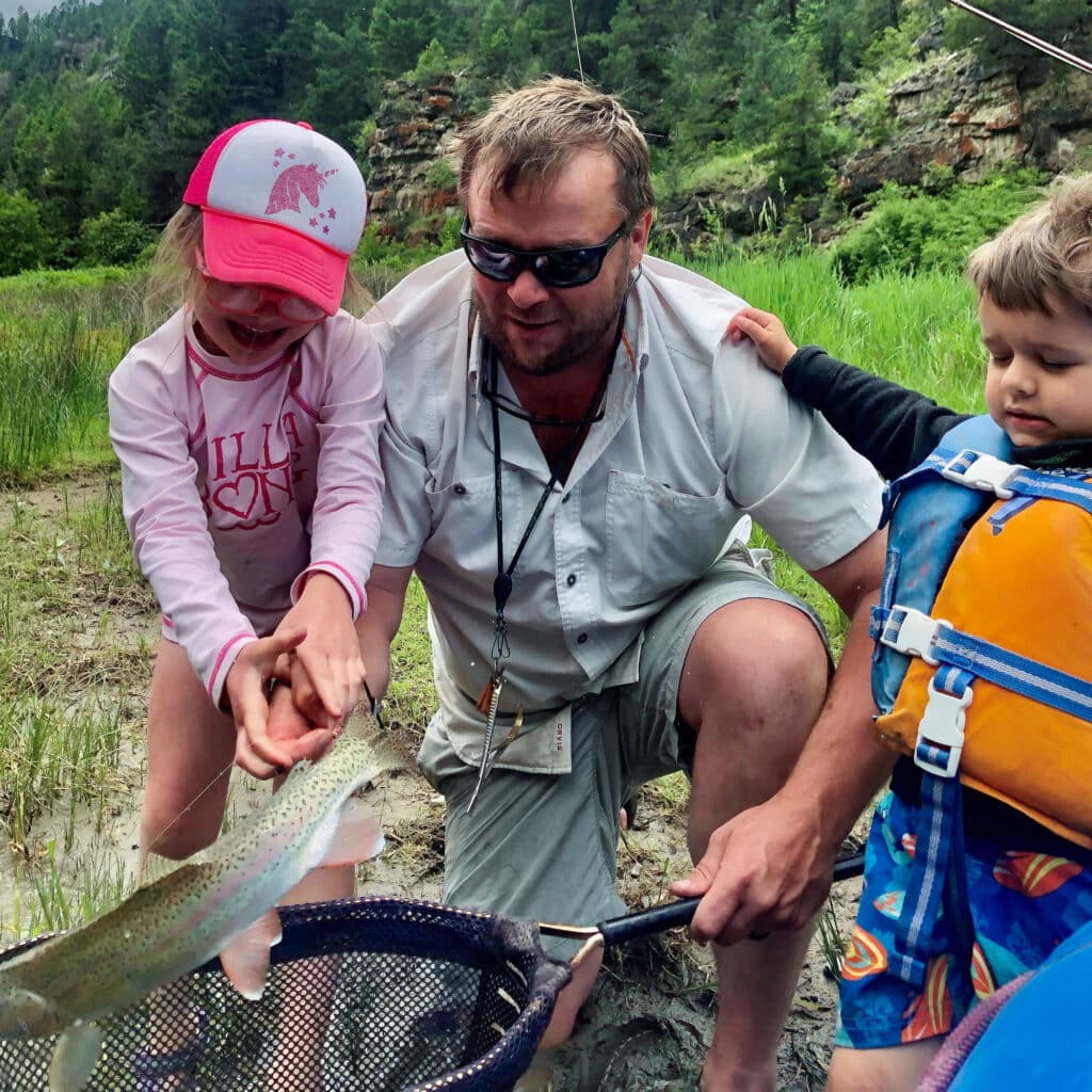 A man kneels beside two children outdoors by a grassy riverbank on a picturesque hunting property. The girl, wearing a pink cap, smiles as she holds a large fish with the man's help. The young boy, in a life jacket, looks at the fish.