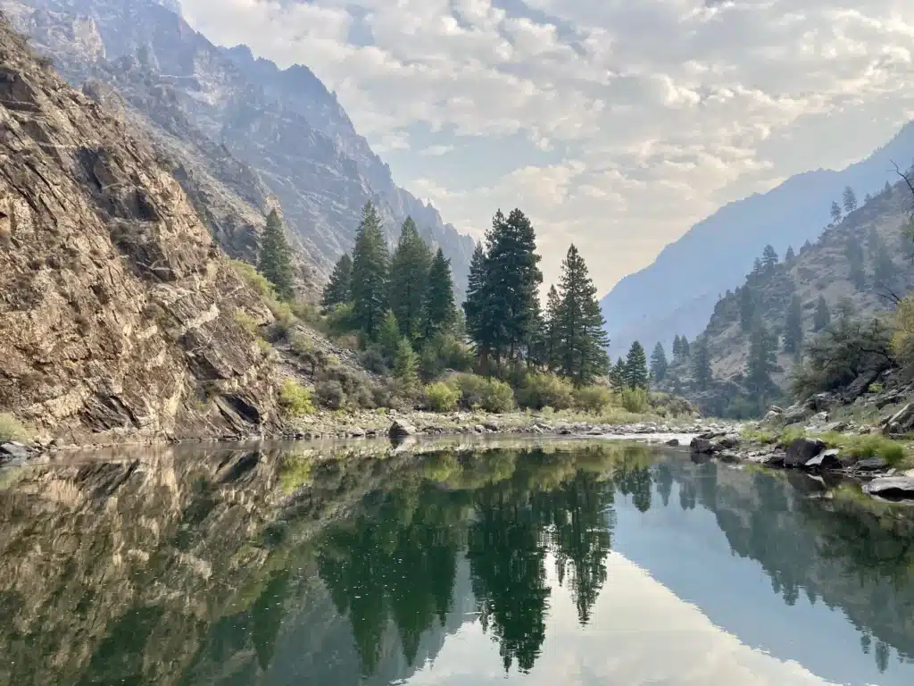 A calm river reflects pine trees and rocky mountains under a partly cloudy sky, creating a mirror-like effect in this serene natural landscape—perfect land for sale for your dream ranch or hunting property.