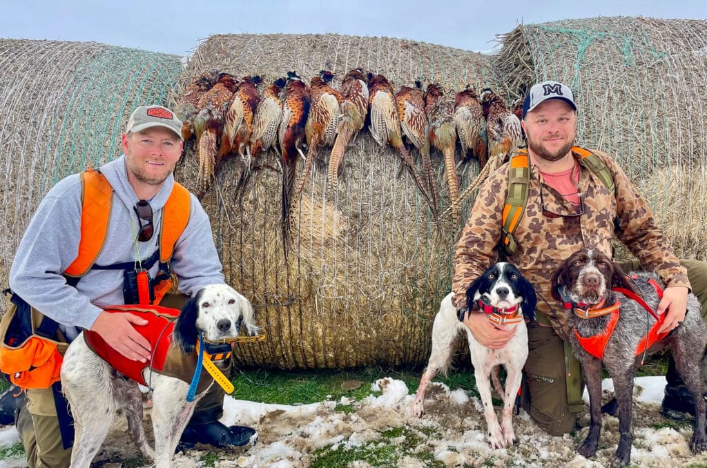 Two men in hunting gear pose in front of hay bales displaying several pheasants. Three hunting dogs wearing orange vests sit on snowy grass, showcasing the possibilities for recreational land or a ranch for sale.