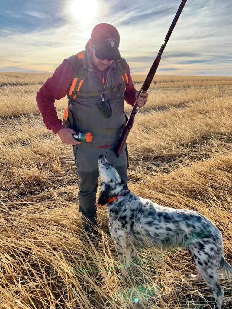 A man in outdoor hunting gear holds a shotgun and a training dummy, standing in a field of dry grass at sunset on recreational land, while a black-and-white dog looks up at him.