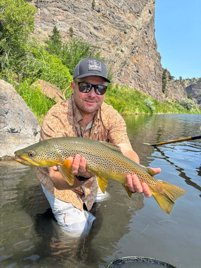A man in sunglasses and a cap kneels in shallow river water on recreational land, proudly holding a large brown trout. Rocky cliffs and green vegetation create a stunning backdrop, perfect for those seeking unique land for sale.