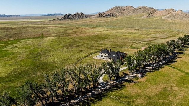 A large house surrounded by trees sits on recreational land in an open, grassy plain with distant rocky hills and mountains under a clear blue sky.