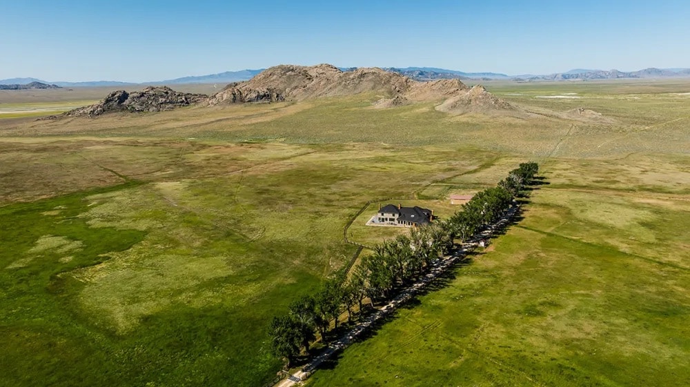 Aerial view of a large green field with a single house surrounded by trees, set near rocky hills under a clear blue sky—ideal recreational land or cattle ranch.