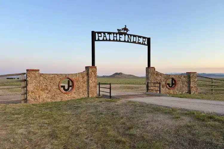 Stone ranch entrance with a metal gate reading PATHFINDER and a cowboy silhouette on top, set in open grassland with distant hills under a clear sky. Perfect hunting property or recreational land, circular JJ brand symbols are on both gate pillars.