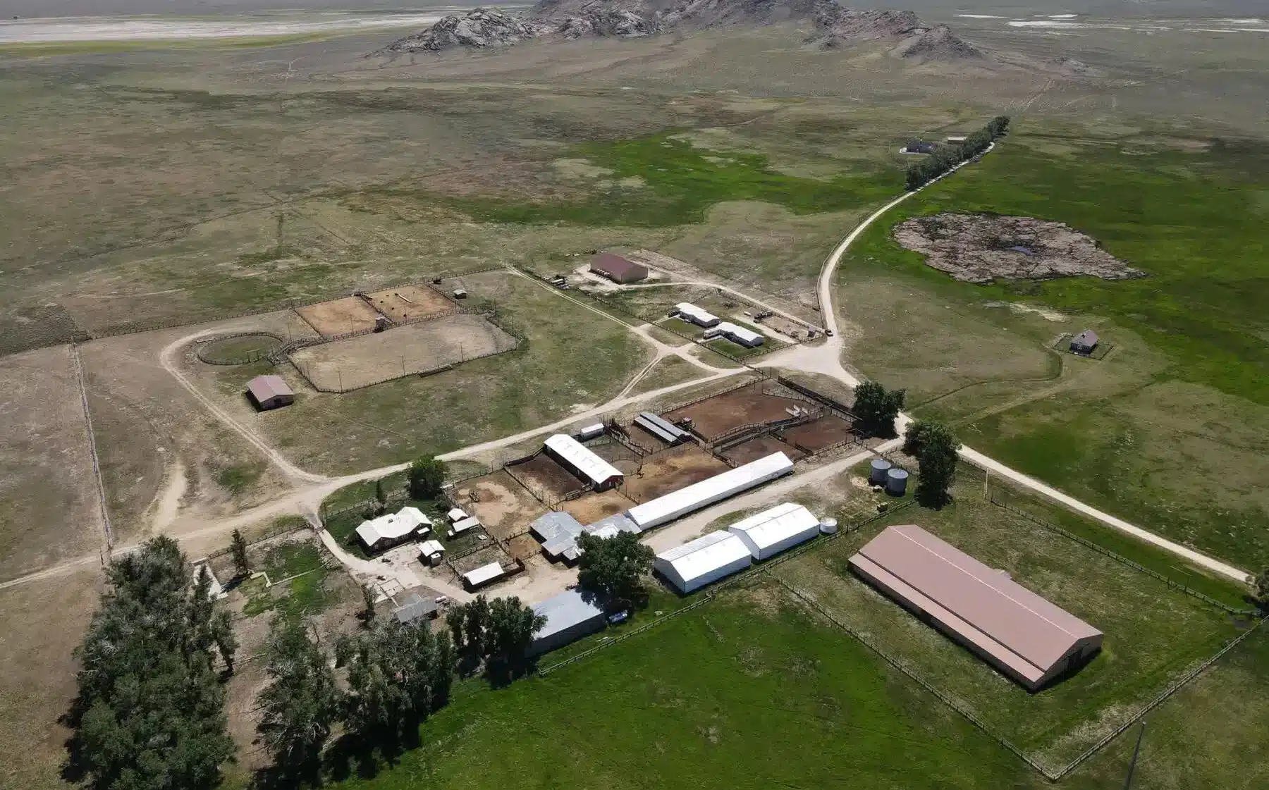 Aerial view of a cattle ranch with barns, outbuildings, fenced areas, dirt roads, and green grass patches surrounded by open fields and distant hills—ideal recreational land.