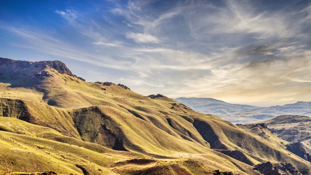 Sunlit rolling hills and rugged mountains under a blue sky with wispy clouds. This picturesque landscape features grassy slopes and distant peaks, perfect for a cattle ranch or prime land for sale, offering vastness and natural beauty.