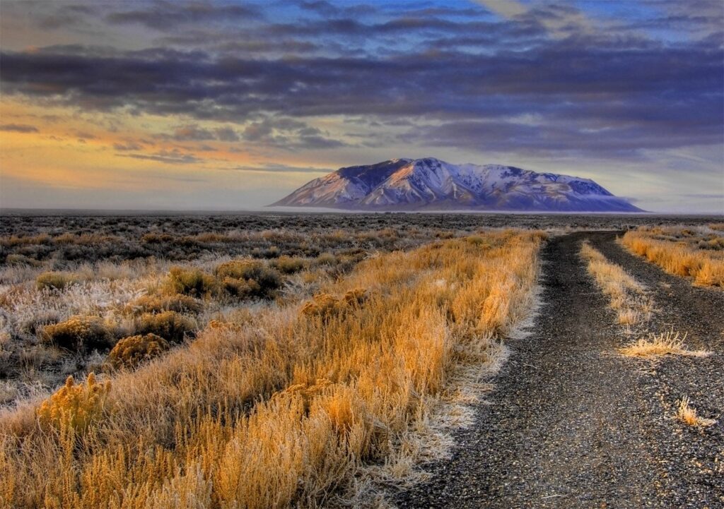 A dirt road winds through dry, grassy plains toward a distant, snow-capped mountain under a dramatic, cloud-filled sunset—perfect land for sale for your dream cattle ranch.
