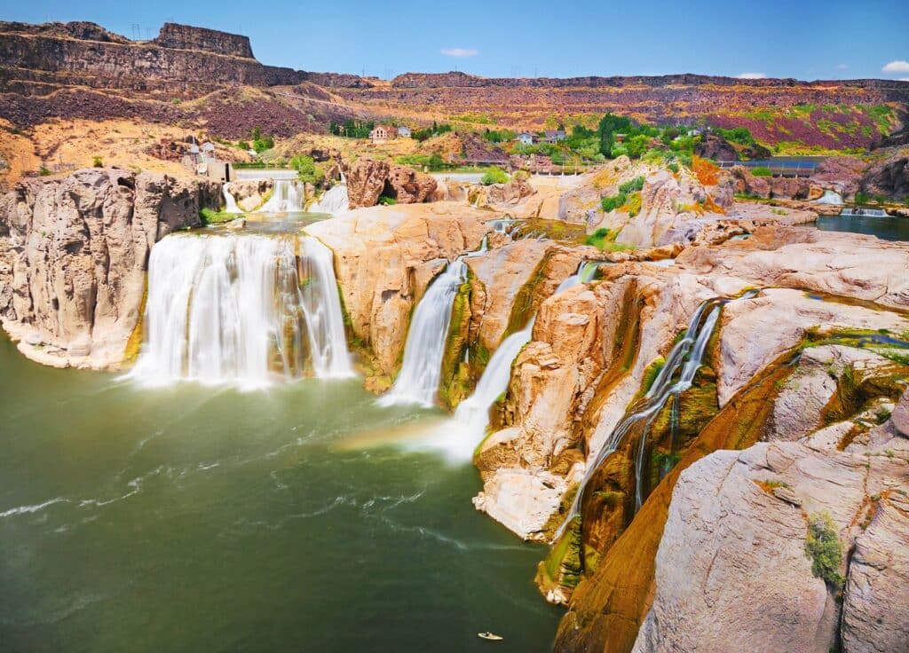 A wide view of Shoshone Falls in Idaho, showing powerful waterfalls cascading over rocky cliffs onto recreational land below, with green vegetation and rugged terrain in the background under a blue sky.