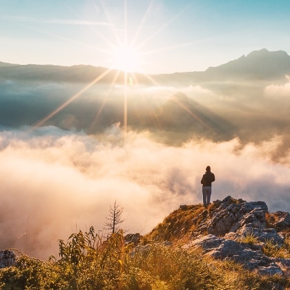 A person stands on a rocky mountain edge above a sea of clouds, facing a bright sunrise with rays stretching over distant mountains—ideal scenery for those seeking recreational land.