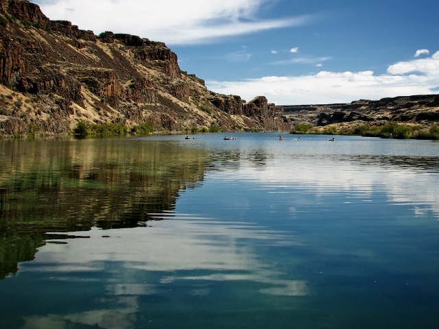 A calm lake reflects rocky cliffs and a blue sky with clouds; several people in kayaks or canoes paddle near the shore, perfect for enjoying recreational land or exploring nearby hunting property.