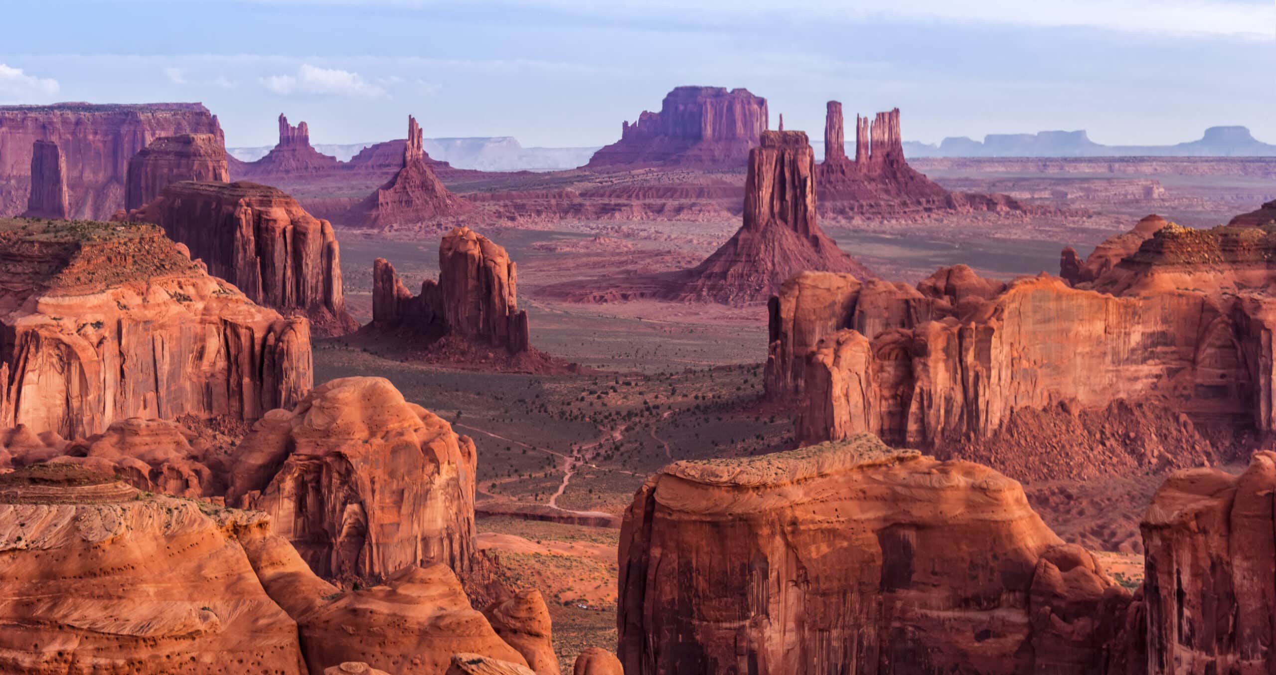 Wide view of Monument Valley with striking red sandstone buttes, mesas, and rock formations rising from a flat desert floor under a partly cloudy sky—an expansive landscape ideal for a ranch for sale or cattle ranch opportunities.