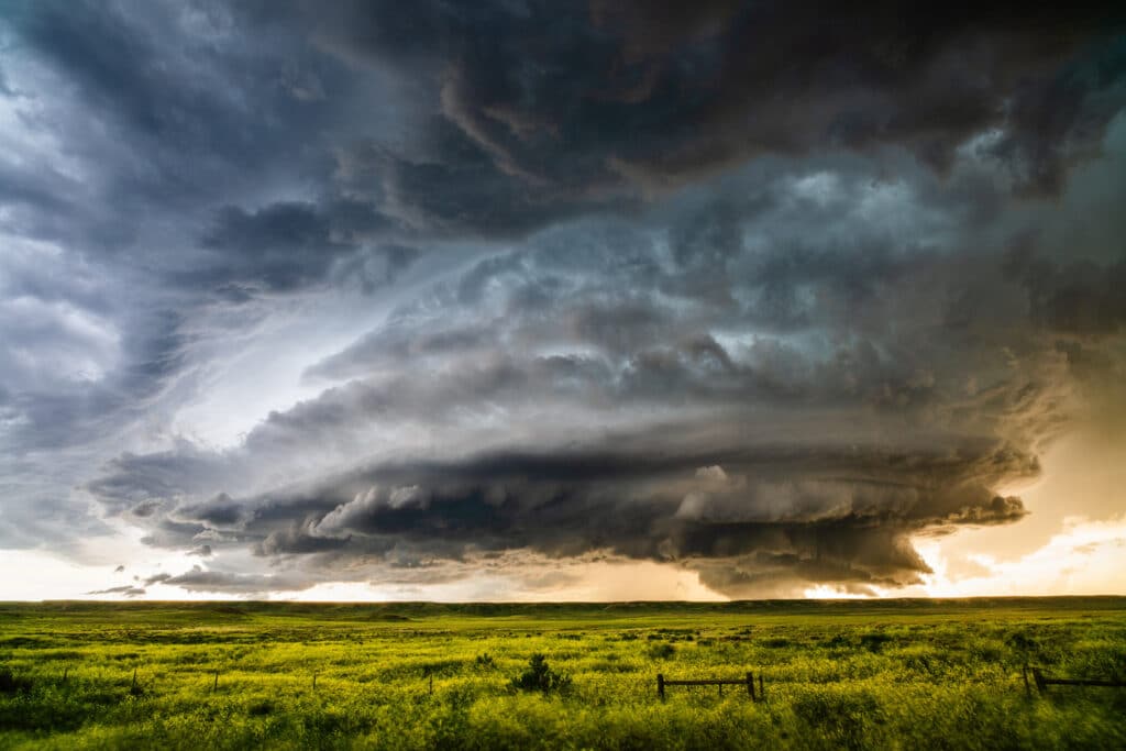 A dramatic, swirling storm cloud hovers over a flat, green field of recreational land. Dark, layered clouds dominate the sky as patches of sunlight break through the horizon, highlighting the striking contrast above this scenic ranch for sale.