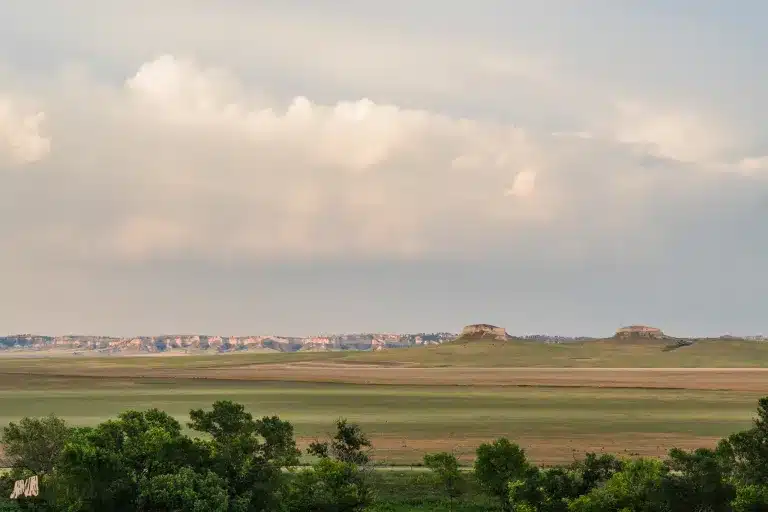 A wide, grassy plain stretches toward distant flat-topped buttes under a mostly cloudy sky, with green trees in the foreground and soft sunlight illuminating this ideal cattle ranch and hunting property.