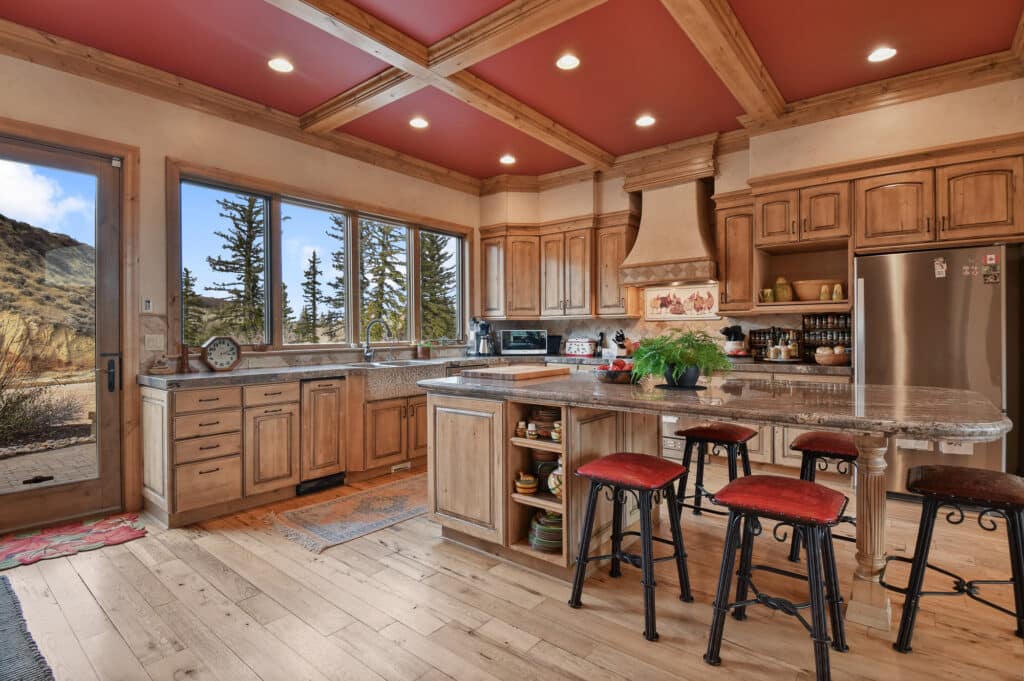 Rustic kitchen with wooden cabinets, red ceiling, stainless steel appliances, and granite countertops. An island with red-cushioned stools sits in the center. Large windows reveal tree-filled views and a mountain landscape fit for a cattle ranch.
