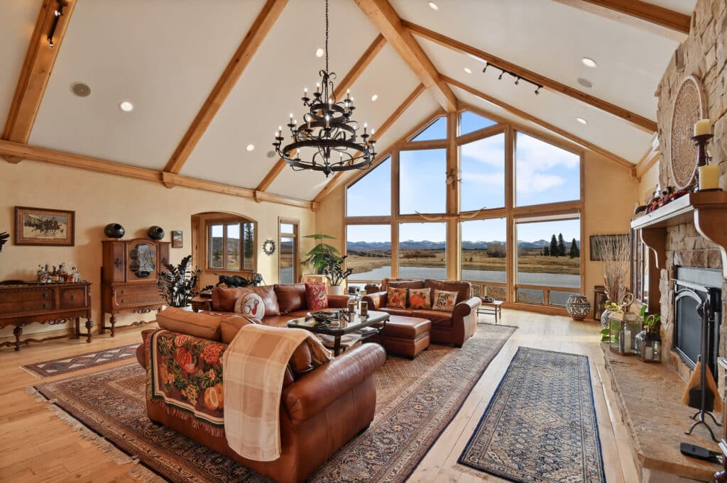 Spacious living room with vaulted ceiling, exposed wooden beams, large triangular windows showcasing scenic recreational land, leather sofas, ornate rugs, a chandelier, and a stone fireplace with decor.