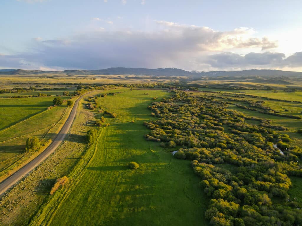 Aerial view of a rural landscape with a winding road, green fields, clusters of trees, and distant rolling hills under a partly cloudy sky in the golden light—ideal recreational land or hunting property for sale.