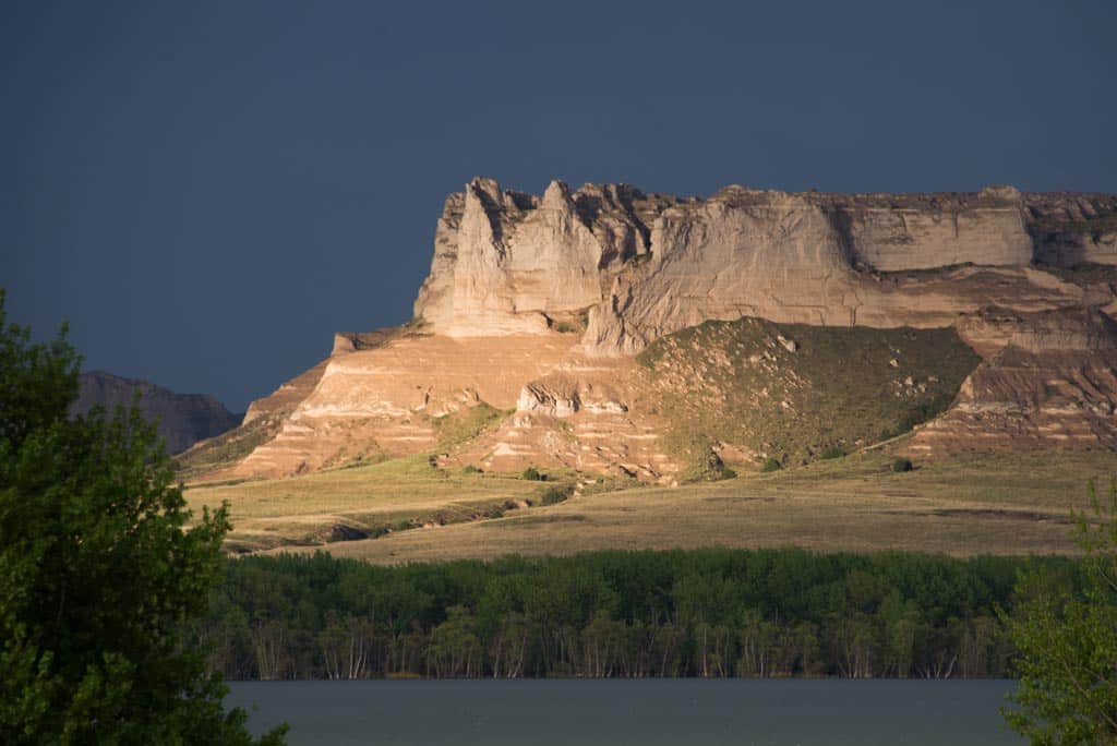 A large, sunlit rock formation rises above a line of green trees and a body of water, set against a dark, cloudy sky—an impressive backdrop for prime land for sale or your future ranch for sale.