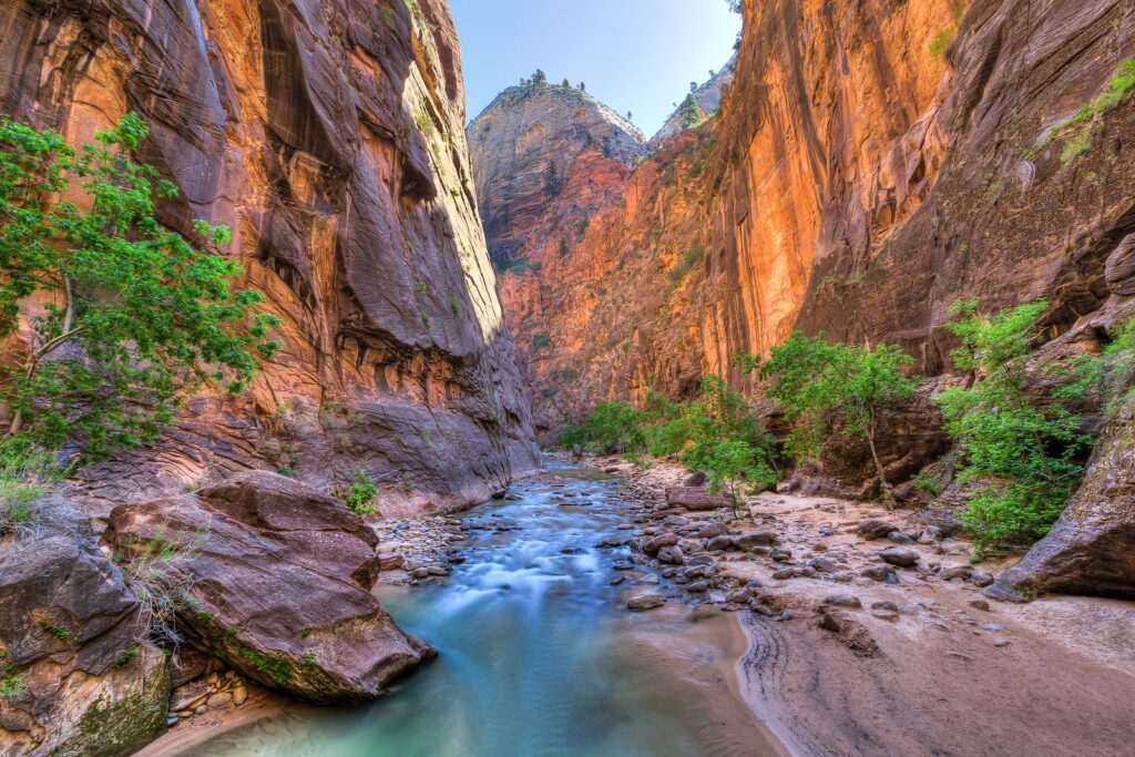 A river flows between tall, steep red rock canyon walls with scattered green trees and shrubs under a clear blue sky, creating a stunning natural landscape ideal for a cattle ranch or hunting property.