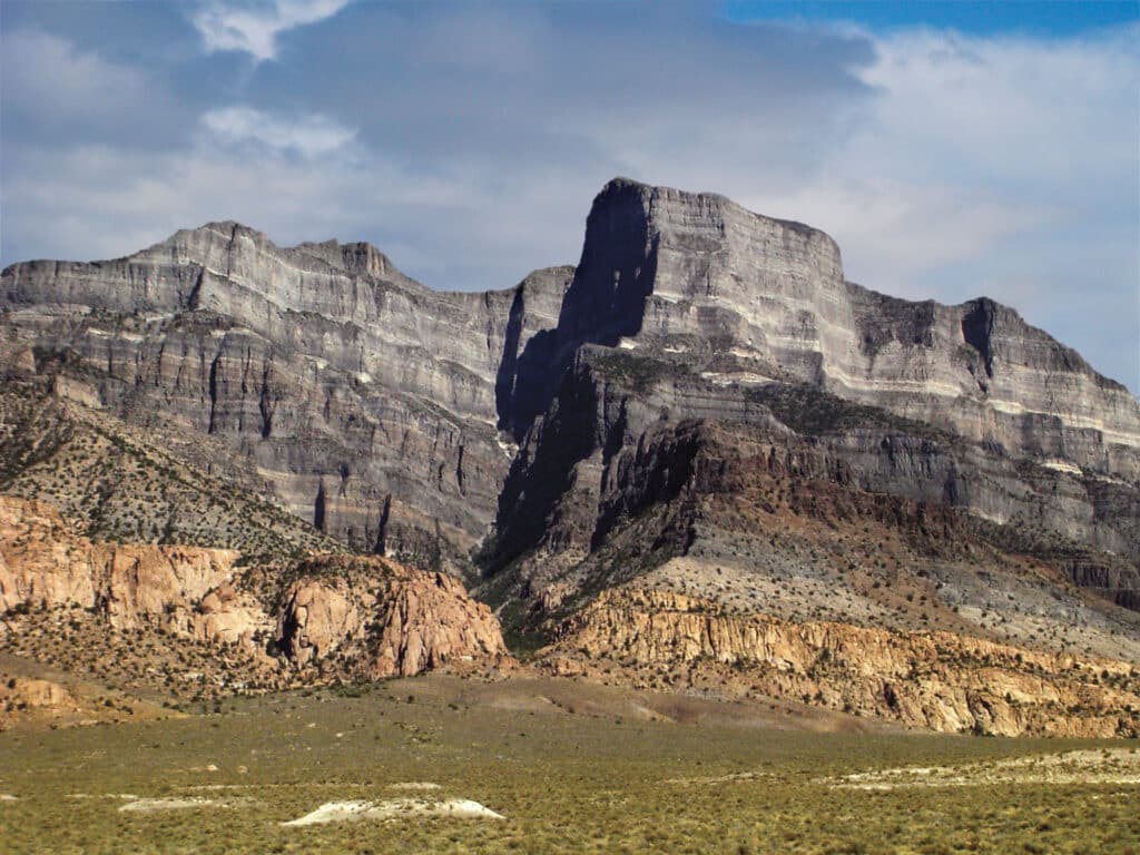 Layered rock formations and rugged cliffs of a desert mountain rise under a partly cloudy sky, with sparse vegetation covering the rocky ground—ideal land for cattle ranch or recreational land enthusiasts.
