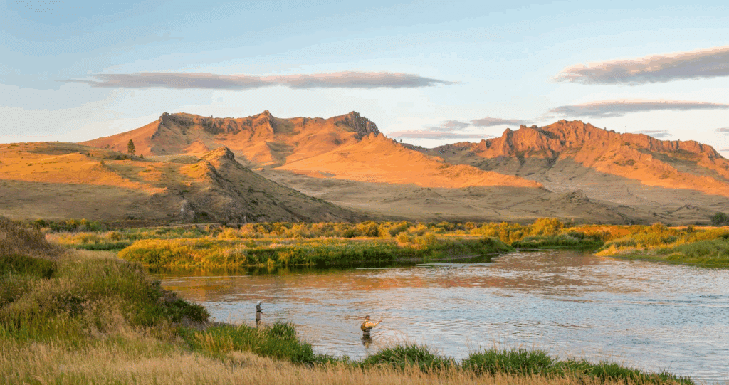 A person fly fishing in a river on recreational land, surrounded by green grass, with rugged mountains in the background illuminated by golden sunlight under a partly cloudy sky.