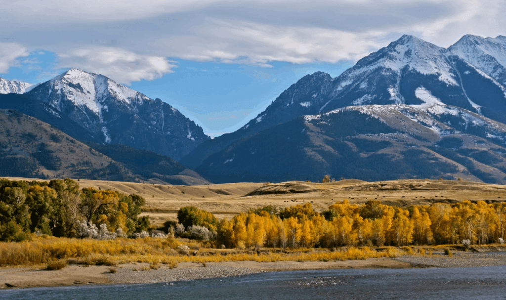 Snow-capped mountains rise behind rolling hills and a line of trees with yellow autumn foliage, with a river in the foreground under a partly cloudy sky—ideal recreational land or hunting property.
