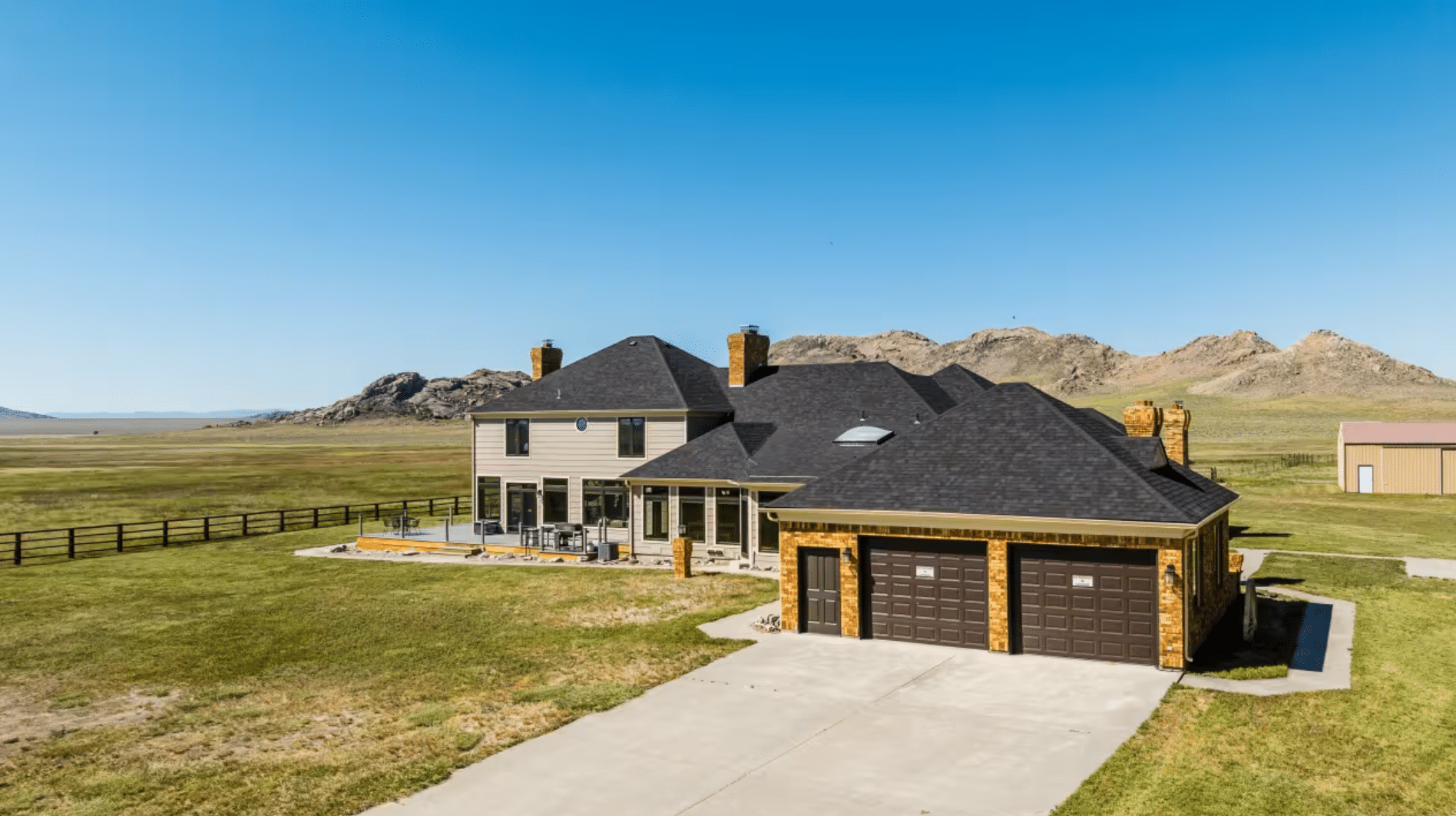 A large house with a dark roof, stone accents, and a three-car garage sits on spacious recreational land, with mountains and a clear blue sky in the background.