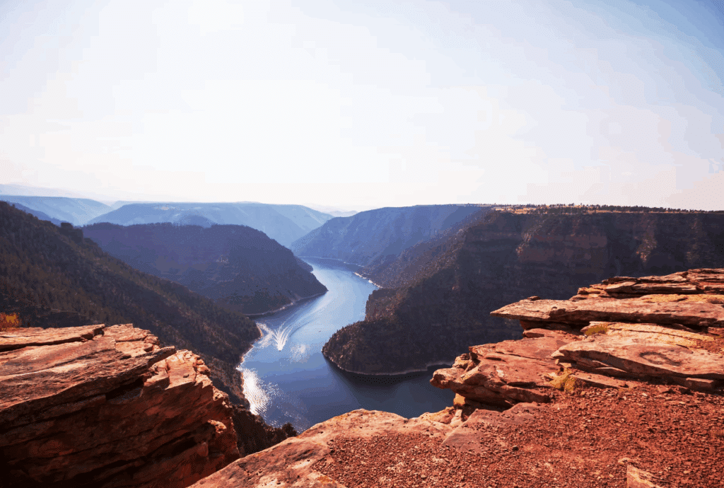 View from a high rocky cliff overlooking a winding river cutting through a deep canyon, with forested hills on both sides under a clear blue sky—ideal hunting property or potential cattle ranch land for sale.