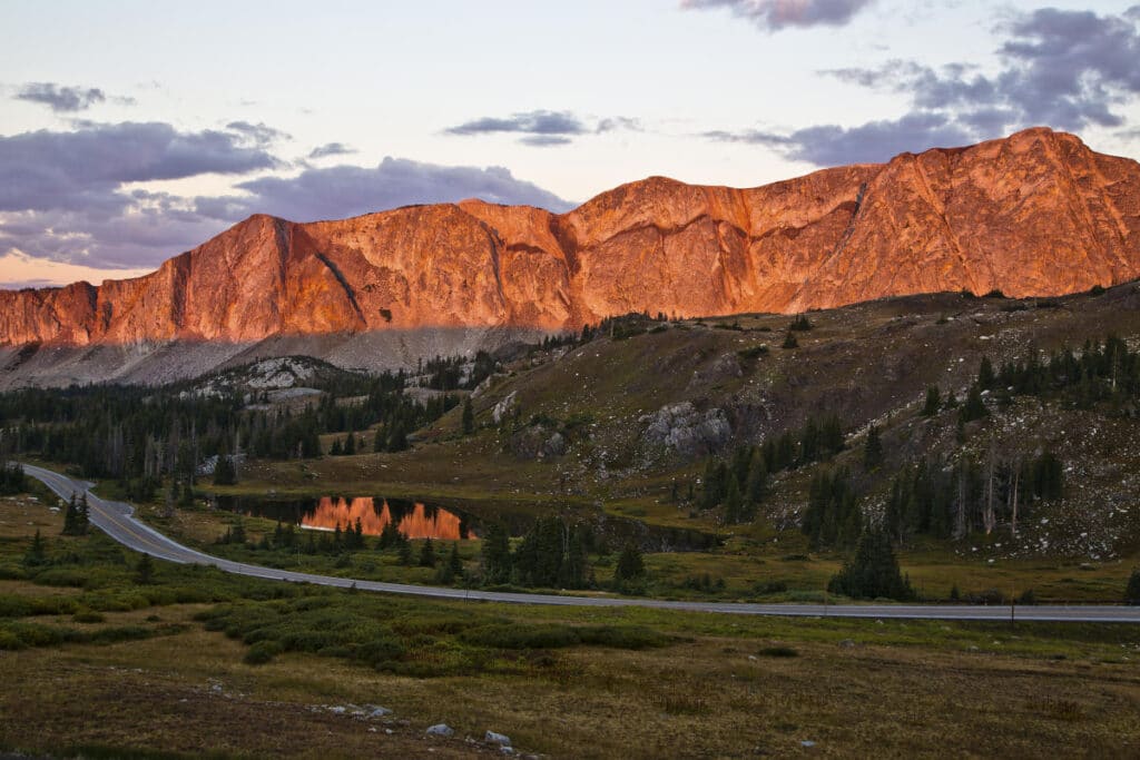 Mountain range glows orange at sunset over a forested valley and winding road, ideal for recreational land. A small pond reflects the bright mountain colors, while scattered clouds hang in the sky.