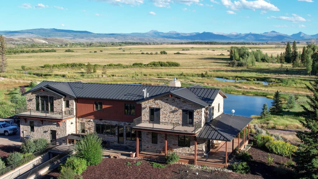 Modern two-story stone house with black metal roof, set in a rural landscape featuring irrigated hay ground and a serene pond, with distant mountains under a blue sky with scattered clouds—a true mountain view retreat and private fishing property in Wyoming.