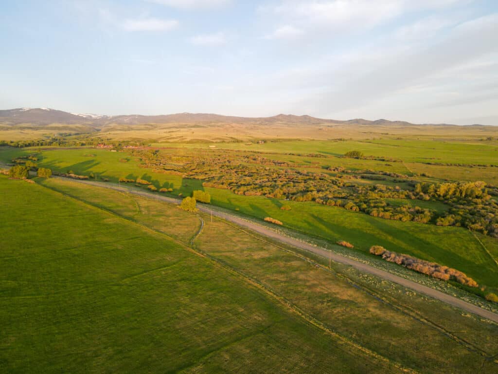 Aerial view of a green countryside landscape with a road winding through fields and patches of trees on a ranch for sale, with distant rolling hills under a partly cloudy sky at sunset.