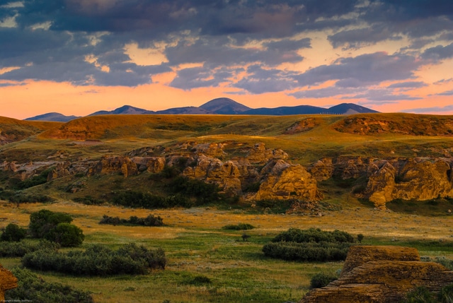 Golden sunlight illuminates rocky formations and grassy plains of this scenic cattle ranch, with rolling hills and distant mountains under a dramatic, partly cloudy sky at sunset.