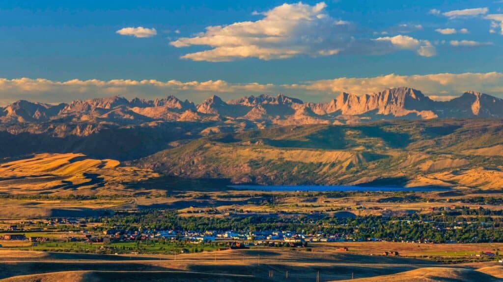 A wide landscape showing a small town surrounded by green fields, perfect as a recreational land, with a lake and rugged mountains in the background under a blue sky with scattered clouds.