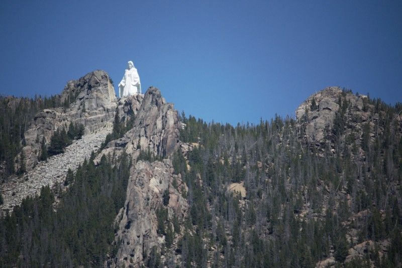 A large white statue of Jesus stands atop a rocky, pine-covered mountain on a scenic cattle ranch under a clear blue sky.