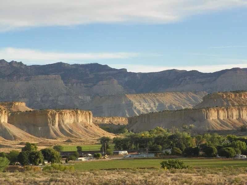 Wide view of a rural landscape with flat-topped, layered sandstone cliffs, green trees, a few scattered houses, and mountains in the distance under a partly cloudy sky—ideal recreational land or ranch for sale.