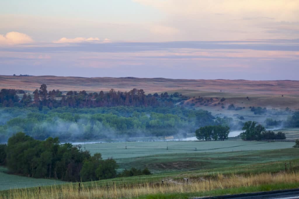 A scenic landscape with rolling hills, green trees, and a river partially covered by morning mist under a pastel-colored sunrise—ideal recreational land or hunting property.