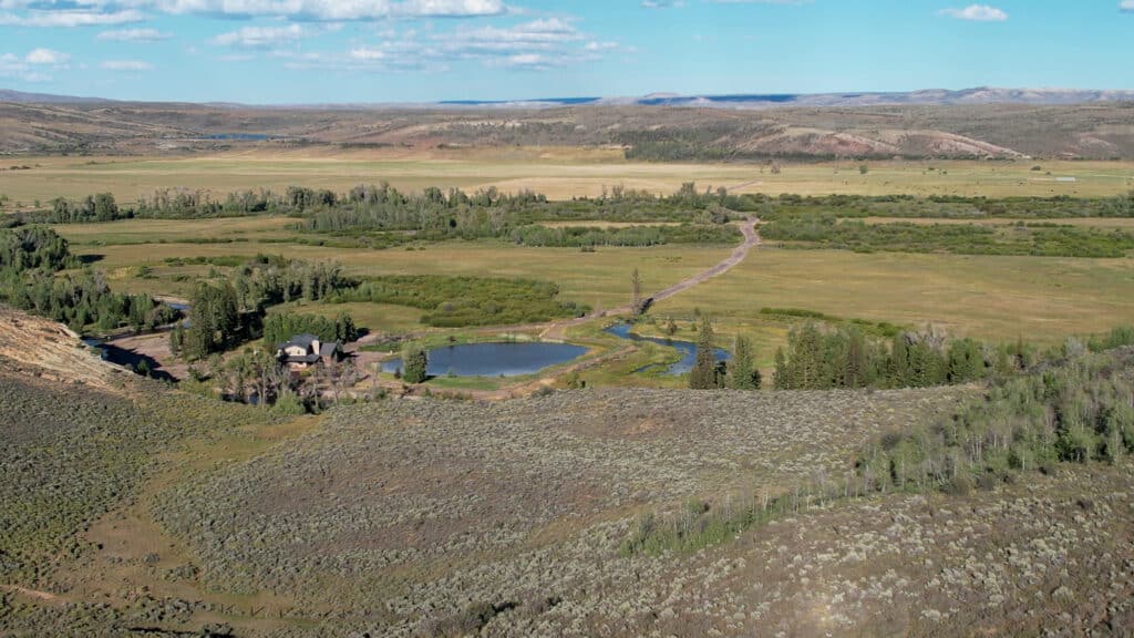 A rural landscape with fields, a winding dirt road, trees, a pond, and a custom home. Distant hills and a partly cloudy blue sky offer a peaceful mountain view in the background.