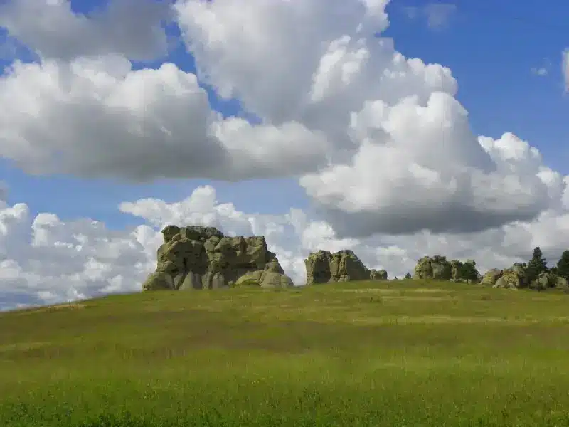 Rock formations rise above a wide, grassy field under a partly cloudy blue sky. This bright and open landscape, with large white clouds casting shadows, is ideal recreational land perfect for those seeking land for sale.