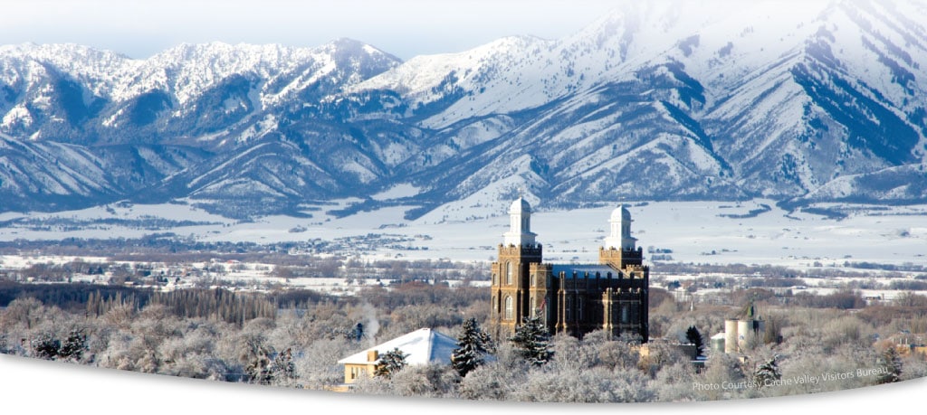 A historic stone building with two towers stands among snow-covered trees on prime recreational land, with snow-capped mountains and a wide, wintry valley in the background under a bright sky.