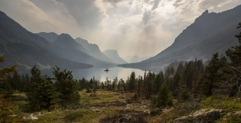 A misty mountain landscape with a calm lake in the center, surrounded by tall peaks and dense evergreen trees under a dramatic sky—ideal recreational land or cattle ranch waiting to be explored.