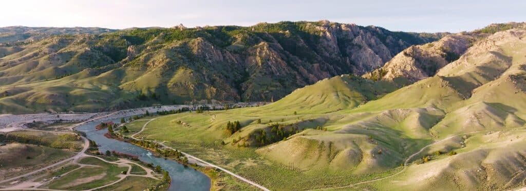 A winding river flows through rolling green hills with rugged, rocky mountains in the background under a clear sky. Dirt roads and sparse trees hint at the untamed beauty of this ranch for sale, ideal as prime hunting property.