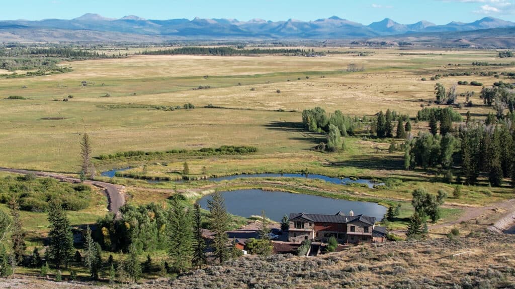 A large Wyoming luxury ranch with a pond and trees sits in a wide, open plain with mountains in the distance under a clear blue sky.