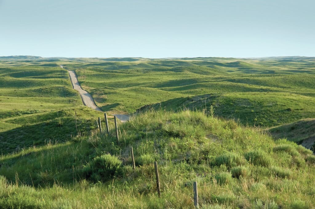 A long, narrow road winds through rolling green hills and grassy fields under a clear blue sky, with a fence running alongside the road—perfect recreational land or hunting property with land for sale in the scenic countryside.