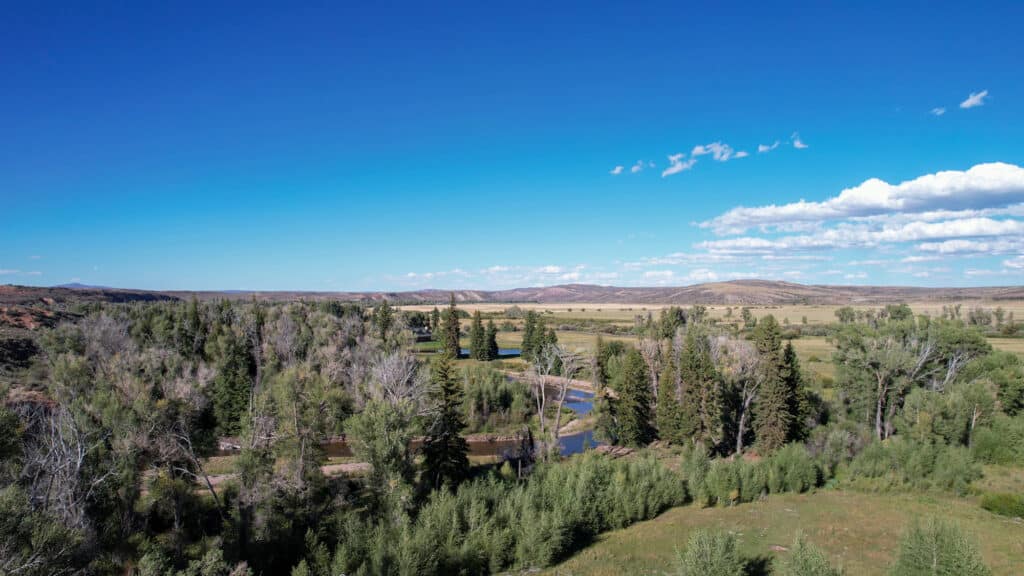 A clear blue sky stretches over a lush green landscape with scattered trees, a winding river, and distant rolling hills under sunlight—perfect for building a custom home with sweeping mountain views and access to irrigated hay ground.