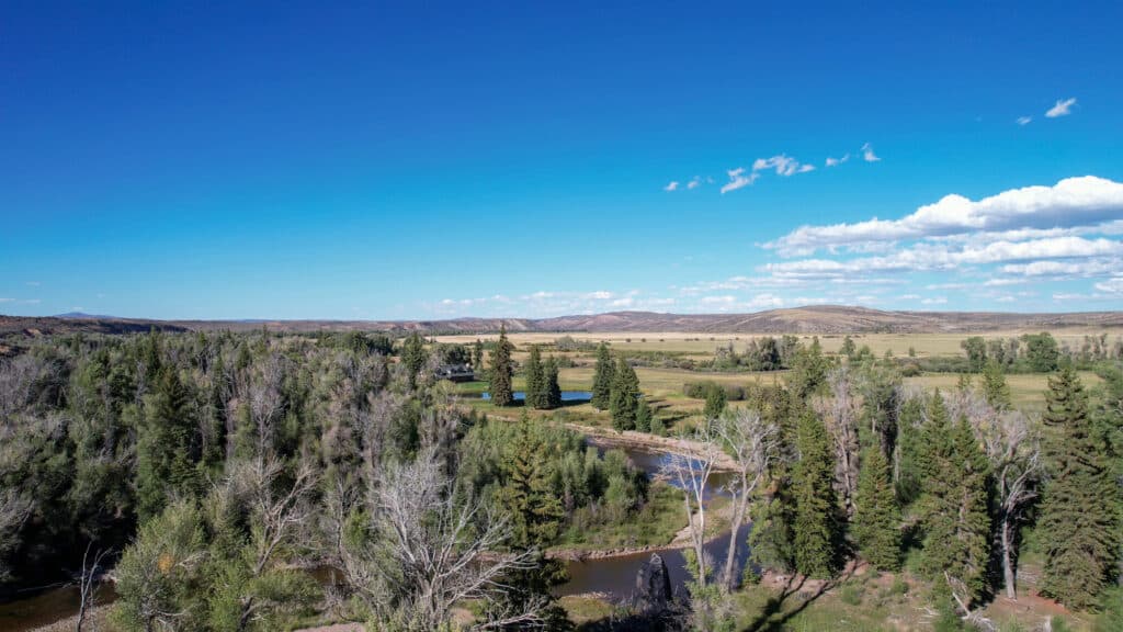 A wide landscape with a river winding through dense green trees, open meadows, and hills under a bright blue sky with scattered clouds—this Wyoming luxury ranch offers breathtaking mountain views and the serenity of private fishing property.