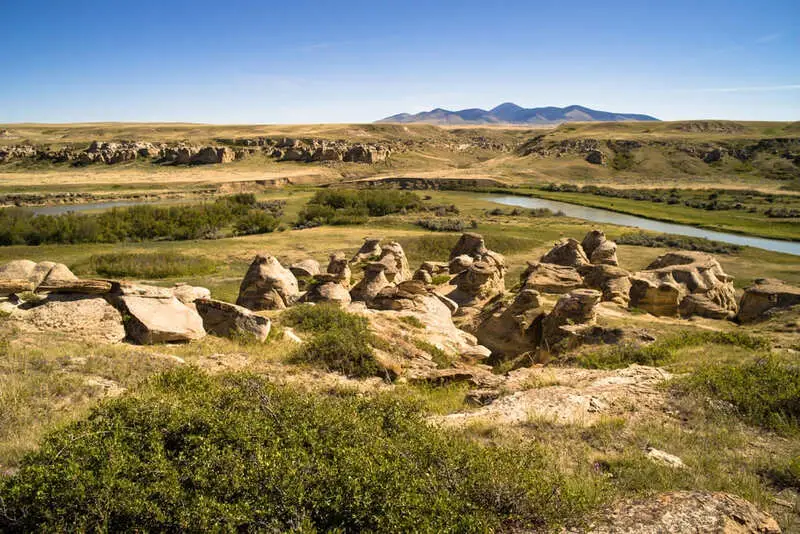 A scenic landscape featuring unique rock formations in the foreground, a winding river, green shrubs, open plains, and distant mountains under a clear blue sky—ideal as recreational land or for use as a cattle ranch.