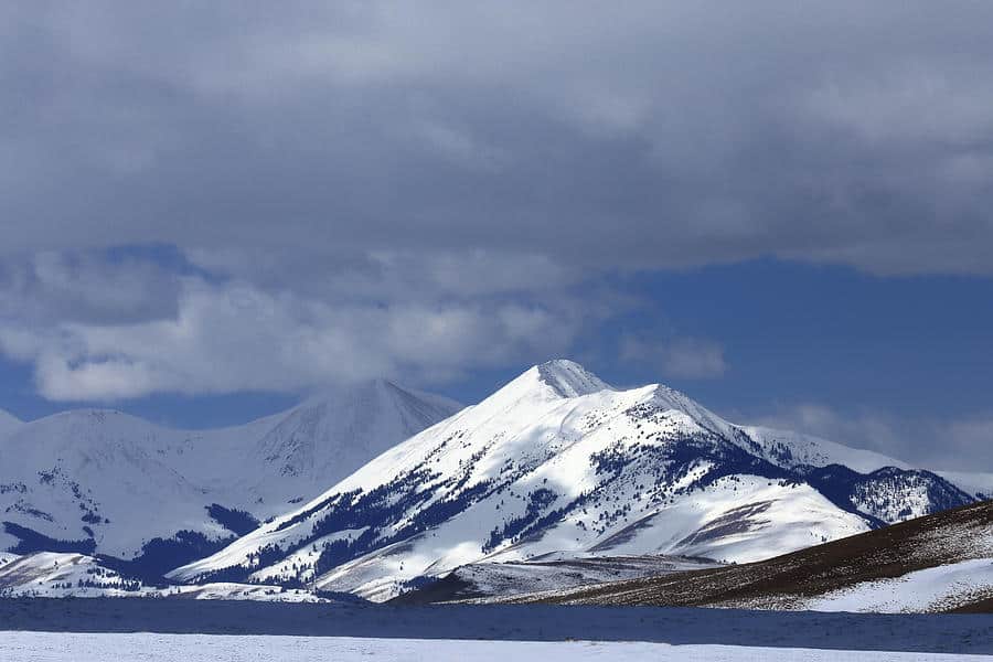 Snow-covered mountains under a partly cloudy sky, with sunlight illuminating the peaks and a stretch of snow-covered recreational land in the foreground—perfect for those seeking a ranch for sale.
