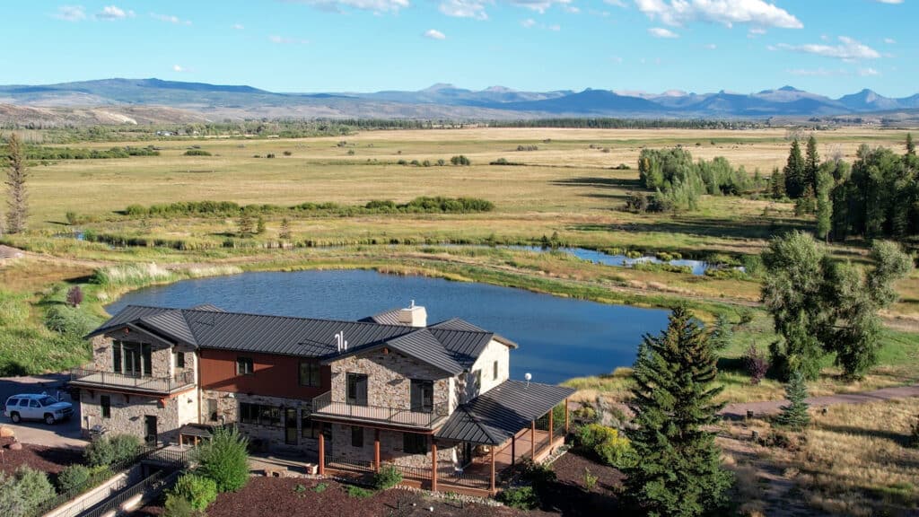 A large stone and wood house sits beside a pond, surrounded by open grassy fields, trees, and distant mountains under a clear blue sky—a true Wyoming luxury ranch featuring irrigated hay ground for lush pastures and endless tranquility.