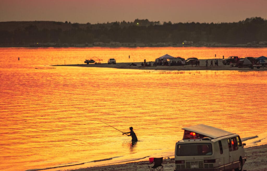 A person stands in shallow water fishing at sunset, with orange reflections on the lake. A white van is parked on the shore of this recreational land, and tents and people are visible on a distant sandy beach.