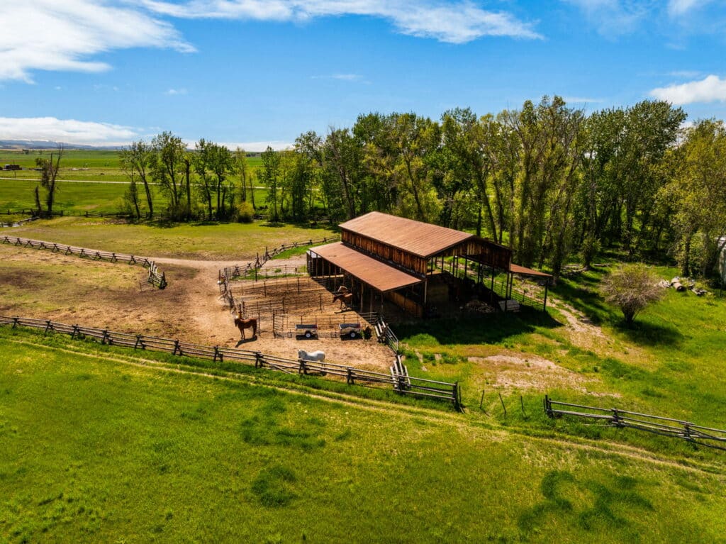 Aerial view of a wooden barn and fenced corral on green grassy recreational land, with one horse standing nearby and trees in the background under a partly cloudy sky. Perfect setting for a cattle ranch or ranch for sale.